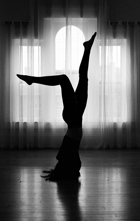 A woman is doing a headstand with her legs in the air, silhouetted against a window with sheer curtains.