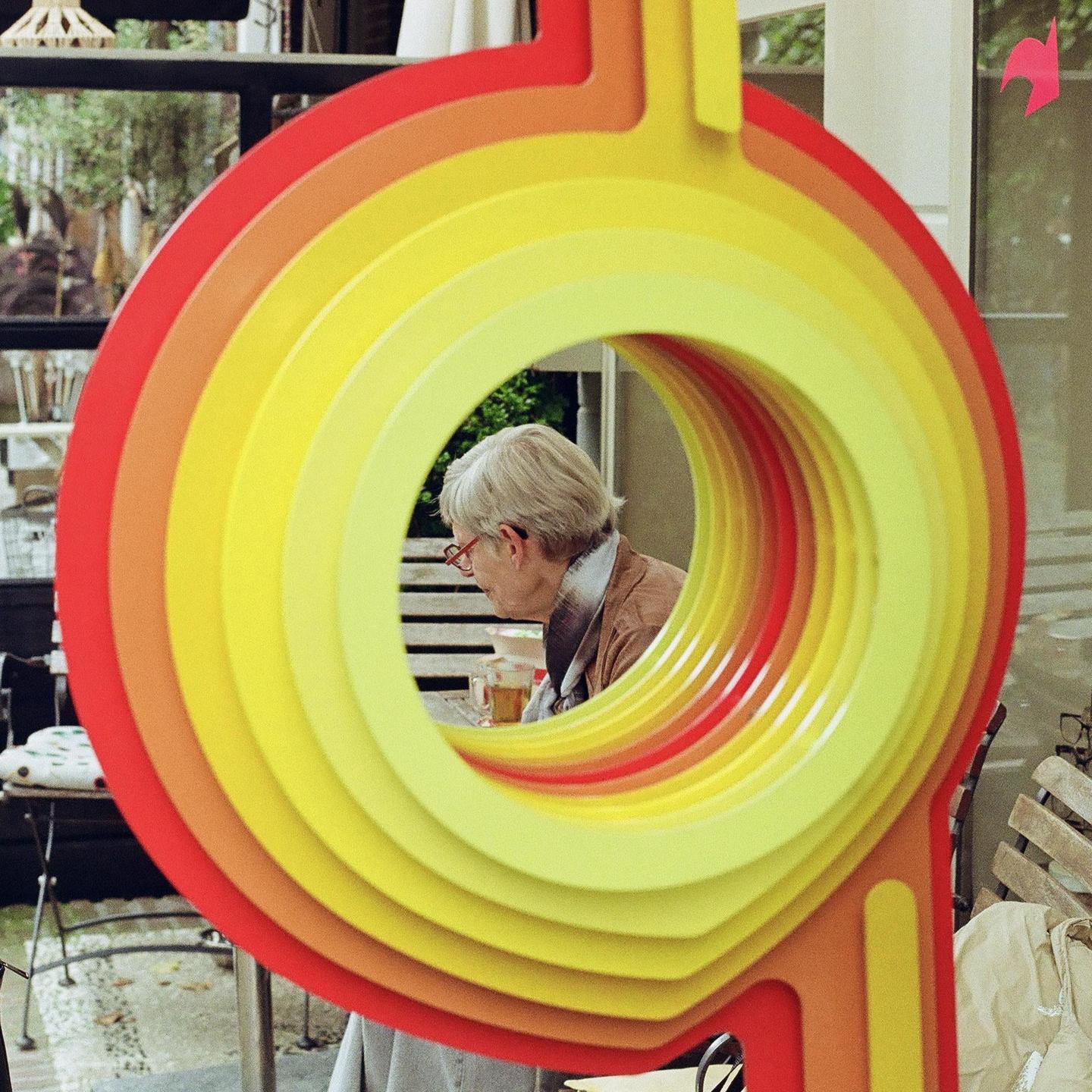A woman with short grey hair and red glasses is visible through a colorful, circular art installation.