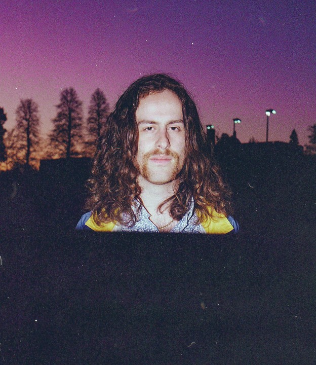 A man with long, curly hair and a mustache looks directly at the camera against a purple dusk sky with silhouetted trees and streetlights.