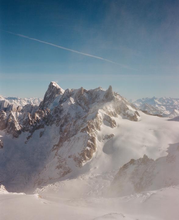 A vast mountain range covered in snow is visible under a clear blue sky with a single contrail.