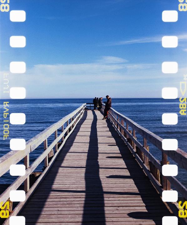 A group of people stand at the end of a long wooden pier stretching into the blue ocean under a clear sky.