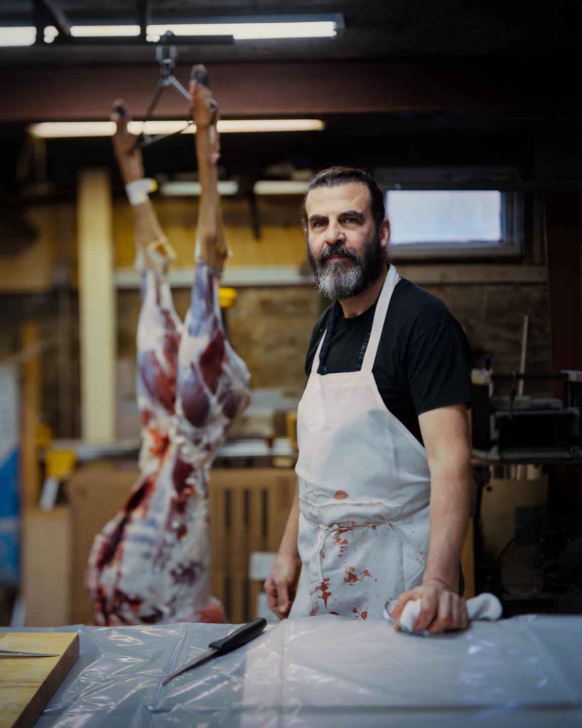 A man wearing a blood-stained apron stands in a garage with a hanging animal carcass in the background.