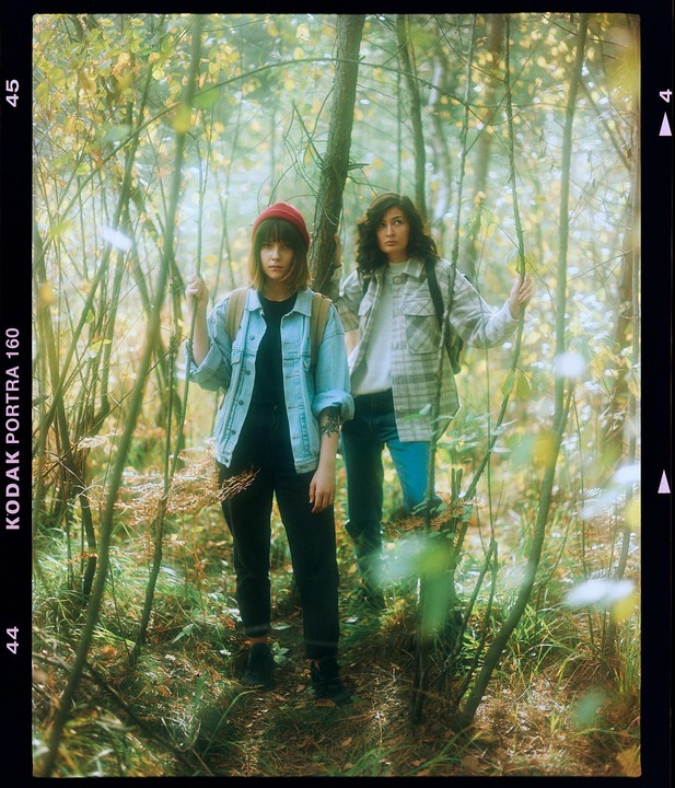 Two women stand among trees and brush in a forest.