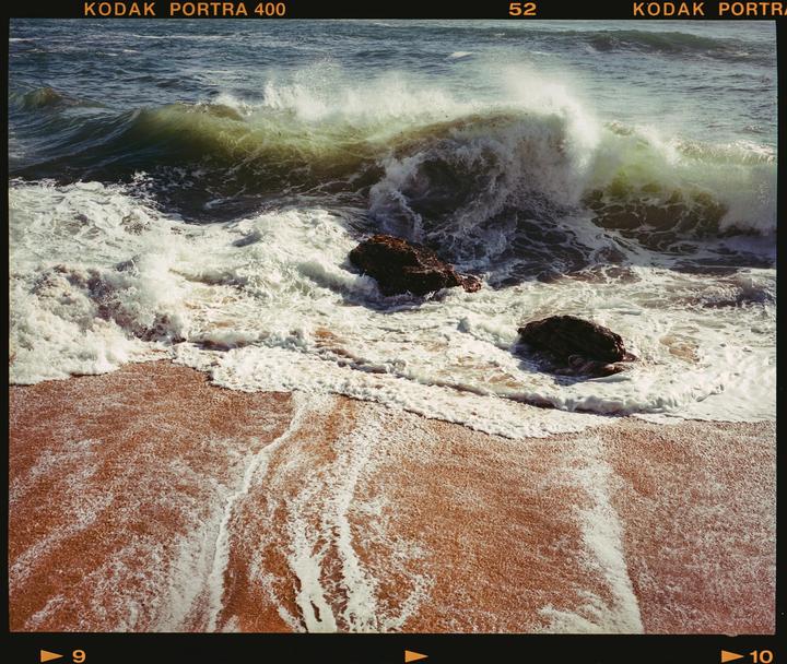 A large wave crashes onto a sandy beach with two rocks visible in the water.