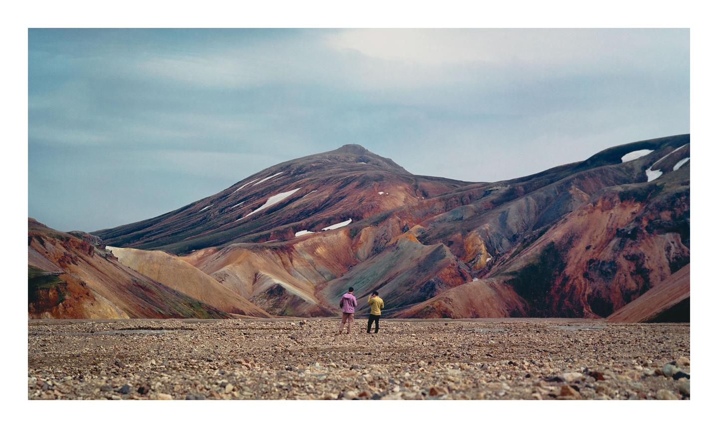 Two people stand on a rocky plain looking towards colorful mountains with patches of snow.