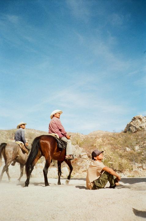 Two men on horseback and one man sitting on the ground are in a desert landscape under a clear sky.