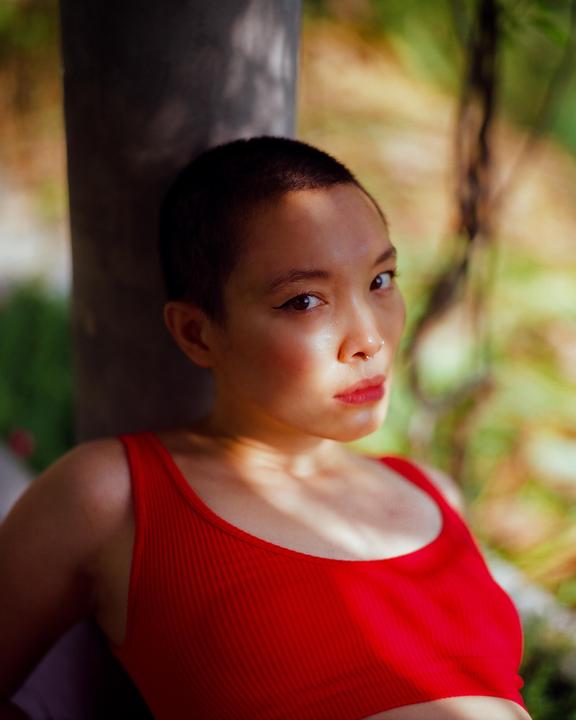 A woman with a shaved head and a red top looks at the viewer while leaning against a tree.