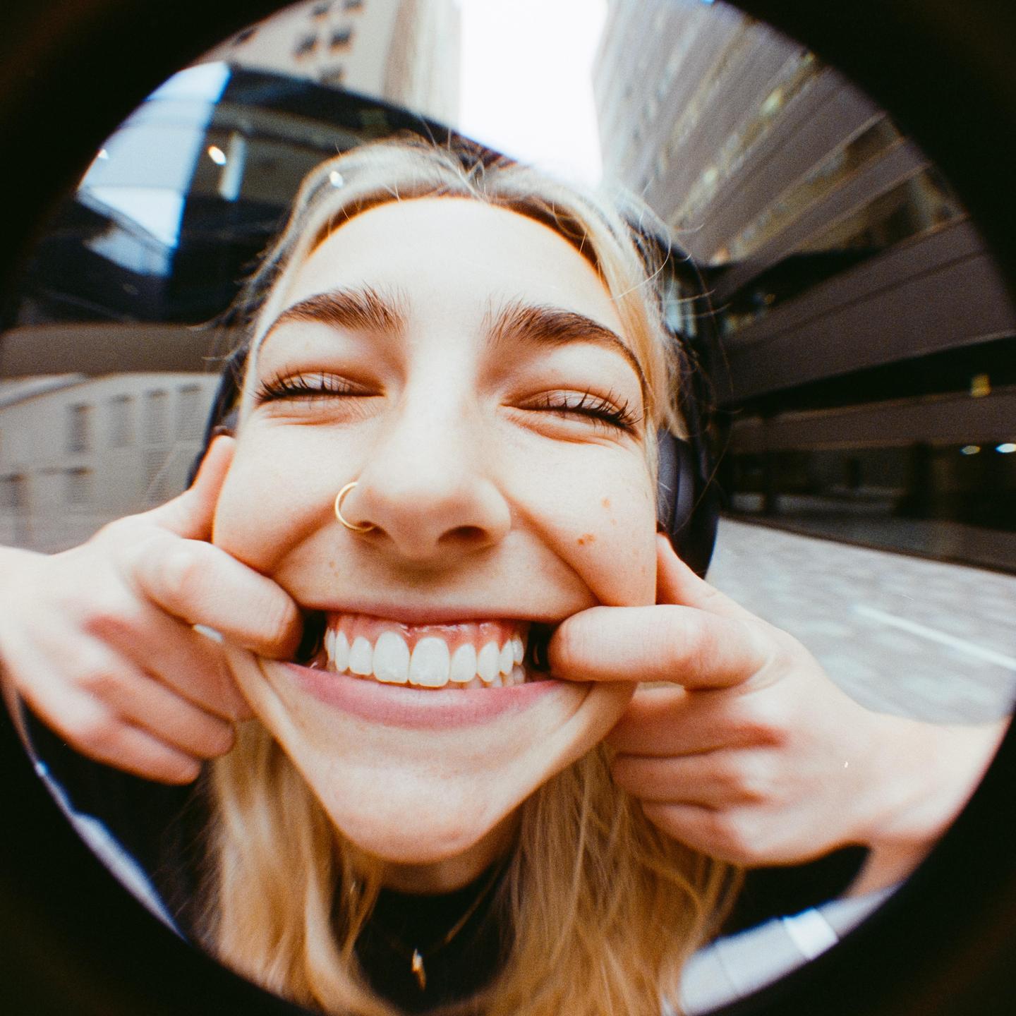 A woman with closed eyes and a nose ring smiles broadly while two hands pull at the corners of her mouth, creating a distorted, wide grin.