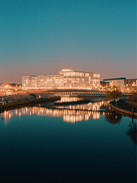 A modern building with many illuminated windows is reflected in the dark water of a river at dusk.