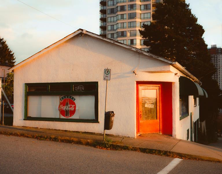 A small white building with a red door and a Coca-Cola sign in the window sits on a sloped street.