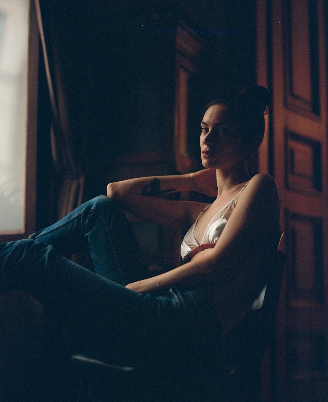 A woman in a white top and jeans sits in a dark room with light coming from a window.