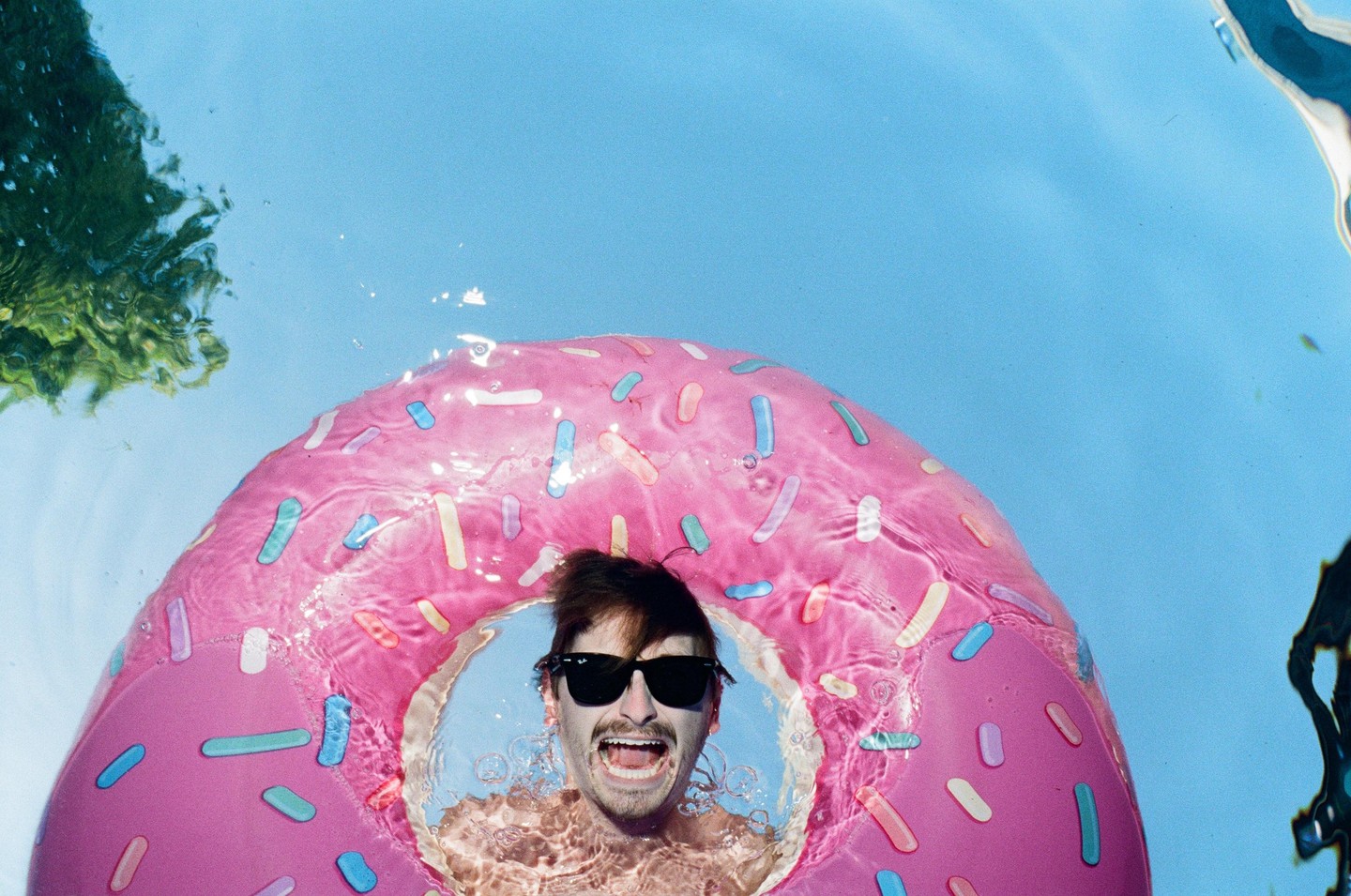 A man wearing sunglasses is in a pink donut-shaped inflatable ring in a swimming pool, with his mouth open.