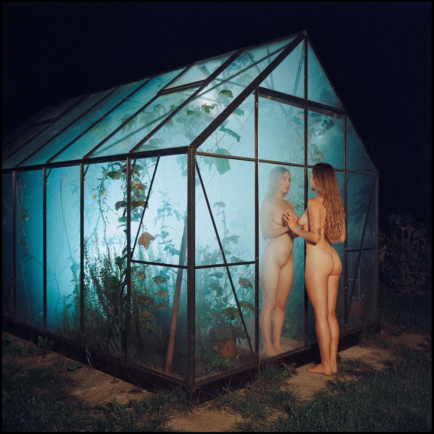 A nude woman stands outside a greenhouse at night, looking at her reflection in the glass.