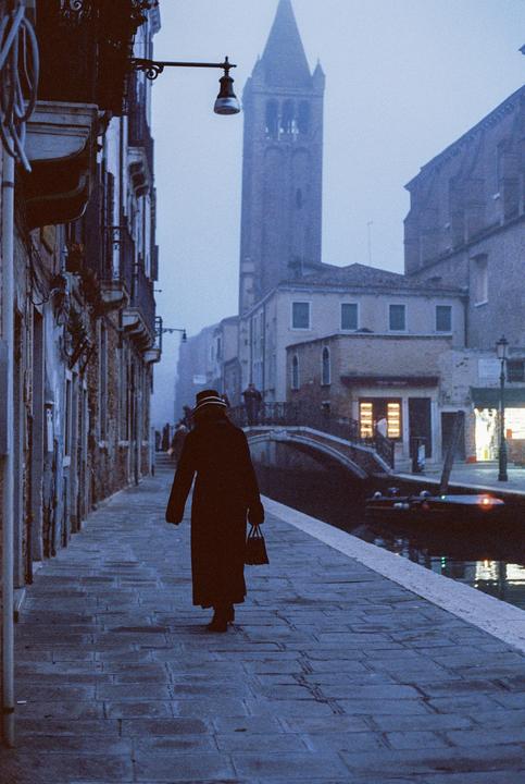 A woman in a long coat and hat walks along a canal in a foggy city with a tall bell tower in the background.