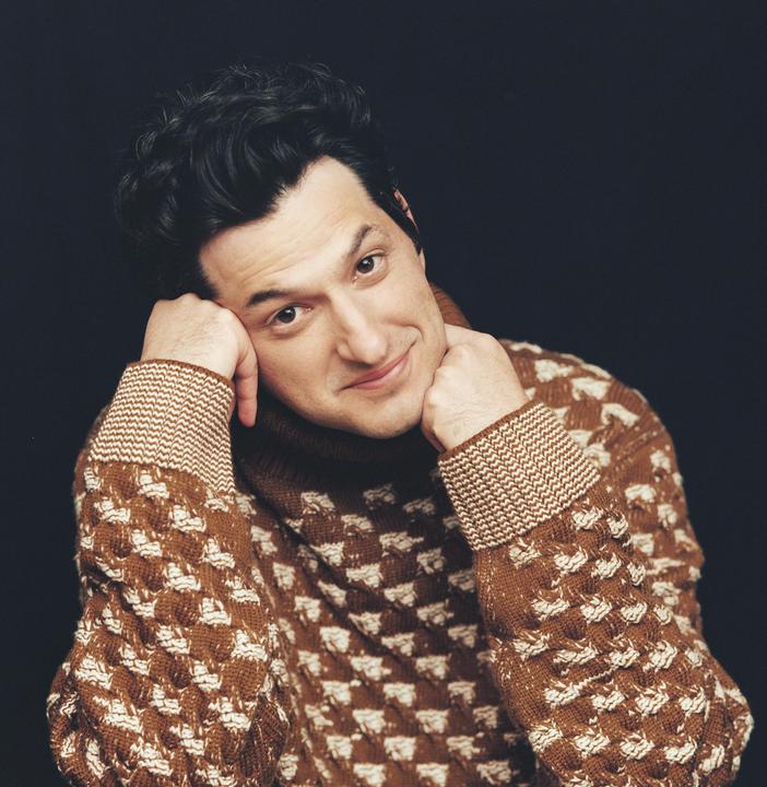A man in a brown patterned sweater rests his chin on his hands and smiles at the viewer.