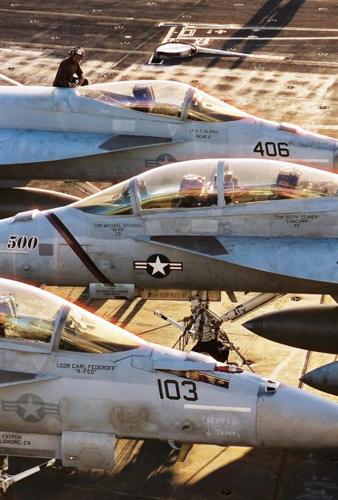 A man sits on top of a fighter jet on an aircraft carrier deck with other jets parked nearby.