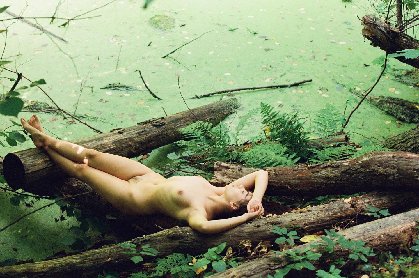 A nude woman lies on logs next to a pond covered in green algae.