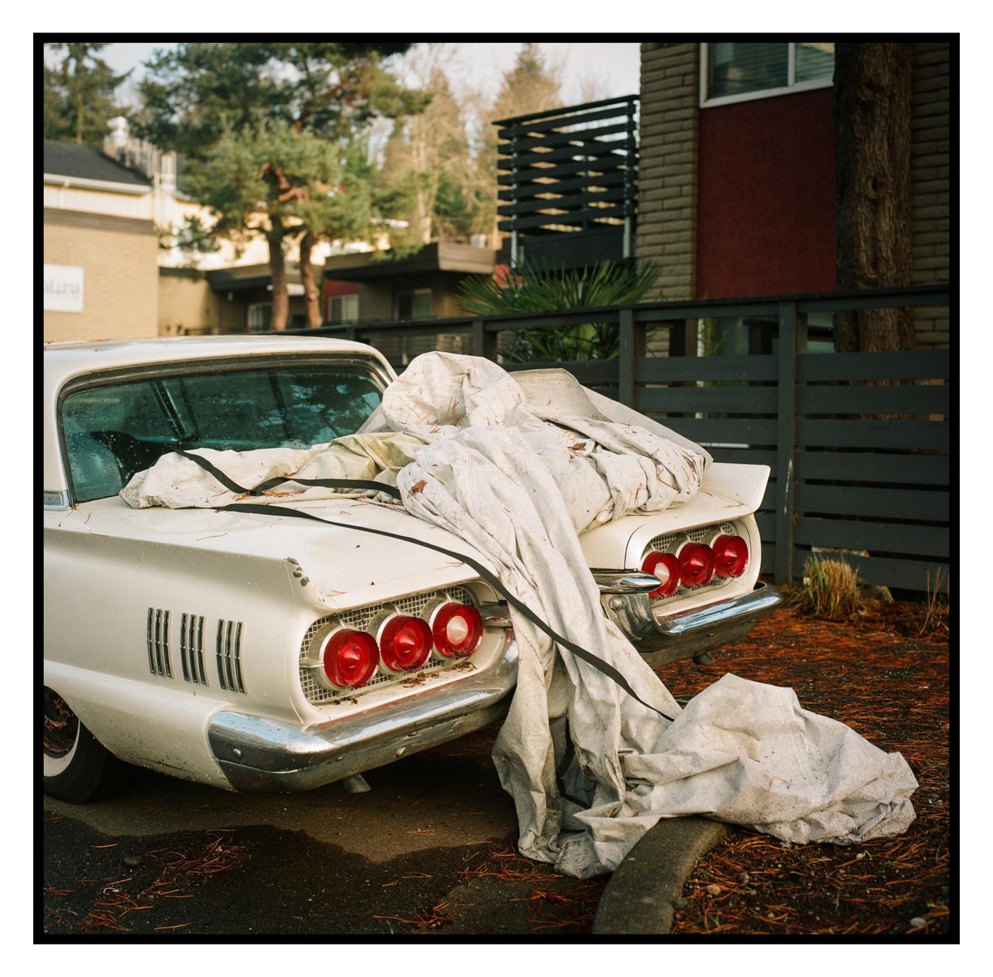 A white vintage car with a white tarp draped over its trunk is parked on a street next to a fence and building.
