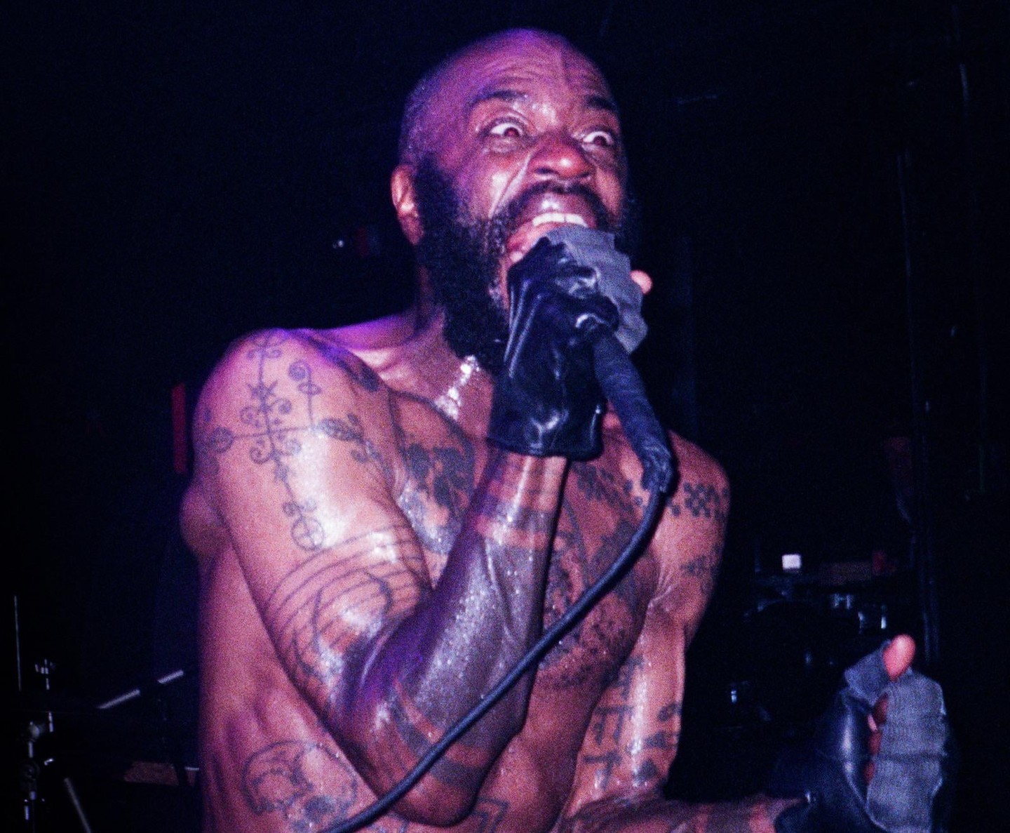 A man with tattoos on his arms and chest holds a microphone to his mouth while performing.