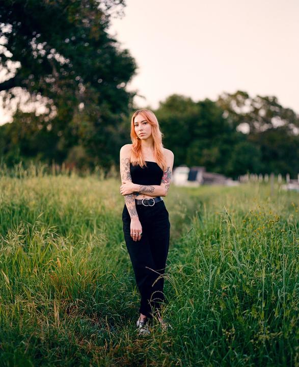 A woman with tattoos stands in a field of tall grass.