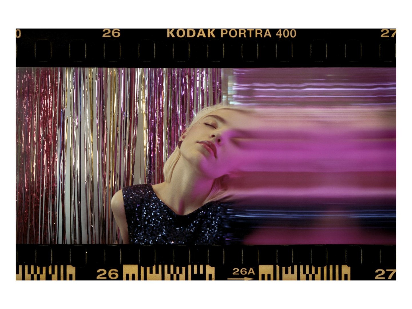 A woman with her eyes closed and her head tilted back is in front of a shiny curtain, with a pink light streak across her face.