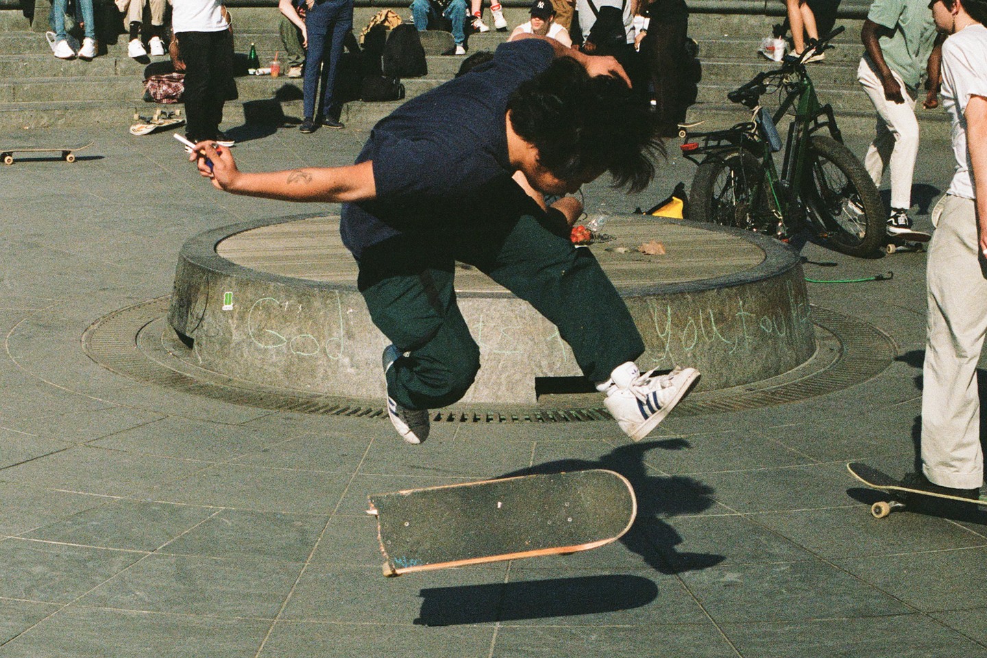 A man in a dark blue shirt and dark green pants is mid-air, jumping off a skateboard on a paved surface.