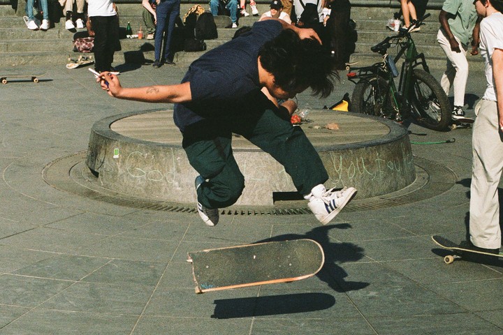 A man in a dark blue shirt and dark green pants is mid-air, jumping off a skateboard on a paved surface.