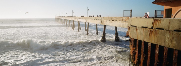 A long pier extends into the ocean with waves crashing below and people walking along its length.