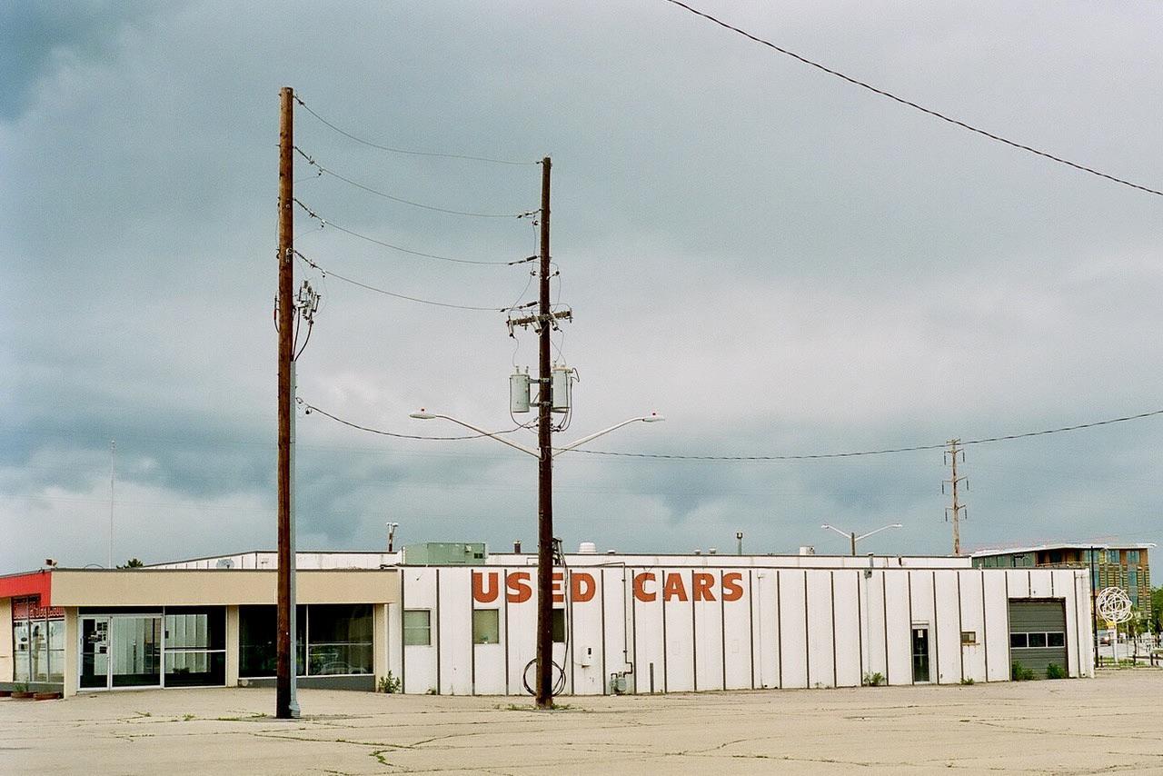 A white building with "USED CARS" written in red letters on its side stands under a cloudy sky, with two utility poles in the foreground.