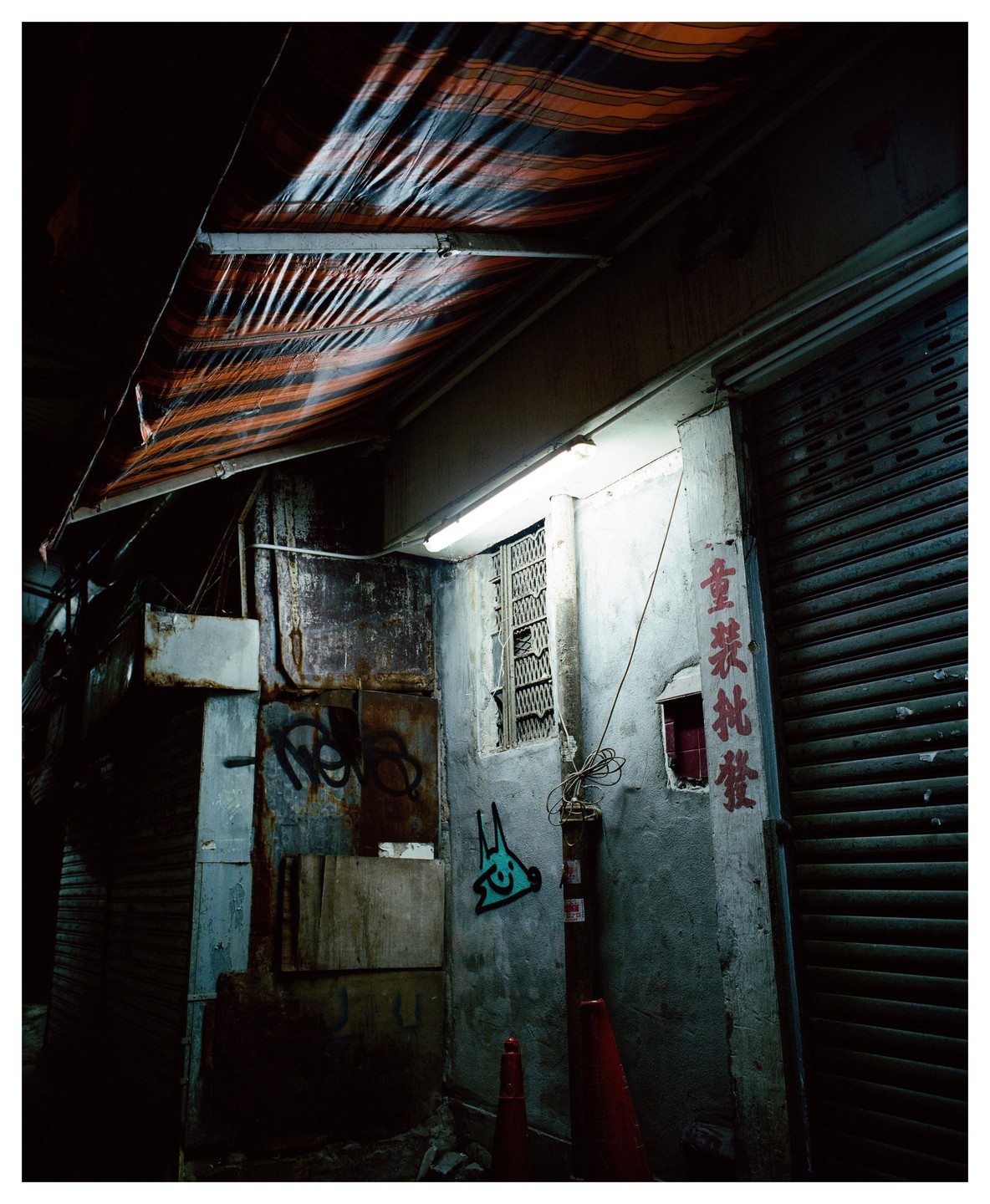 A dark alley in Hong Kong is illuminated by a fluorescent light and features graffiti and a striped awning.
