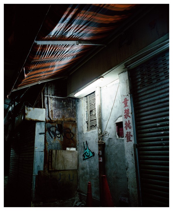 A dark alley in Hong Kong is illuminated by a fluorescent light and features graffiti and a striped awning.
