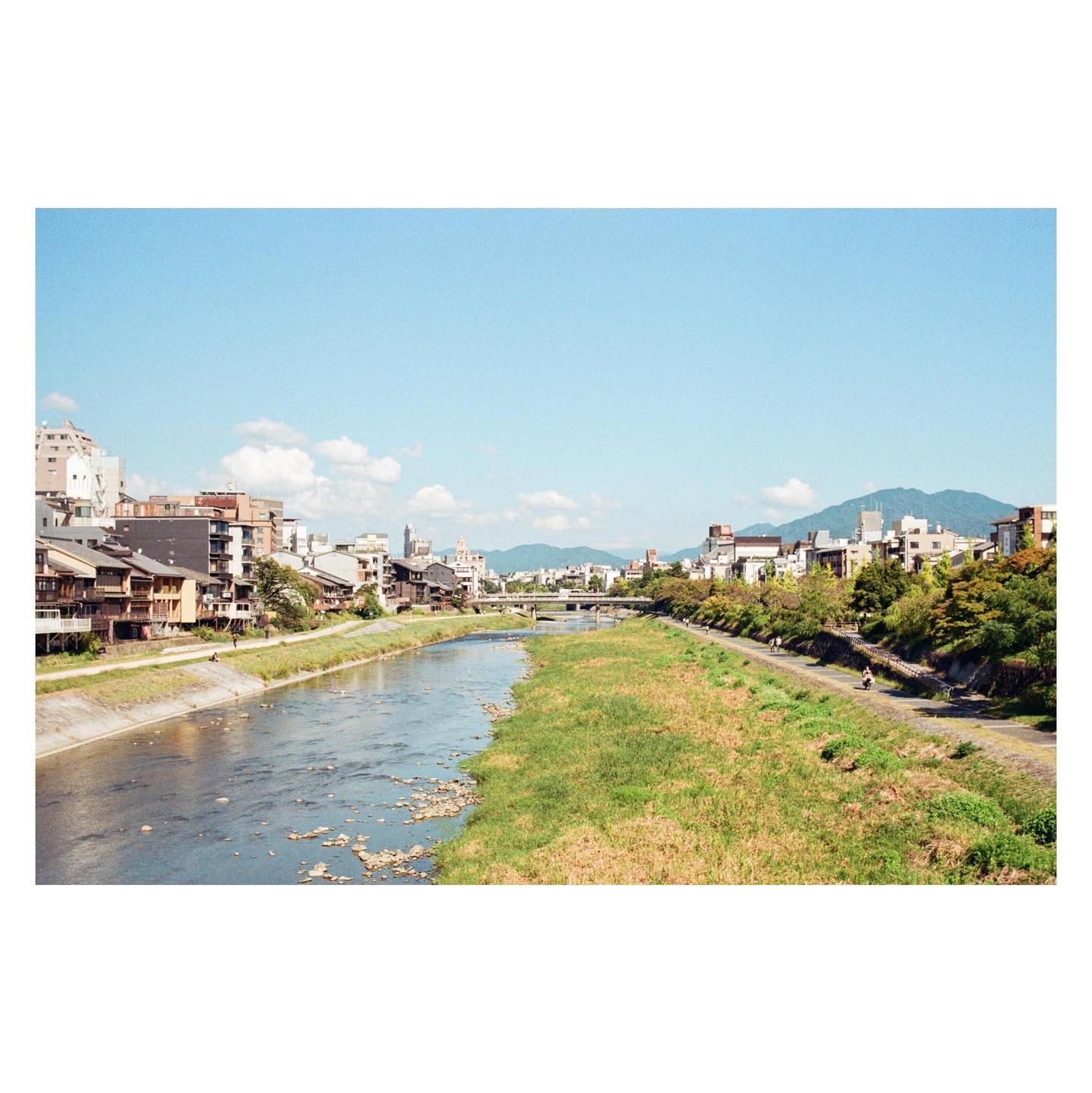 A river flows through a city with buildings and mountains in the background under a clear sky.