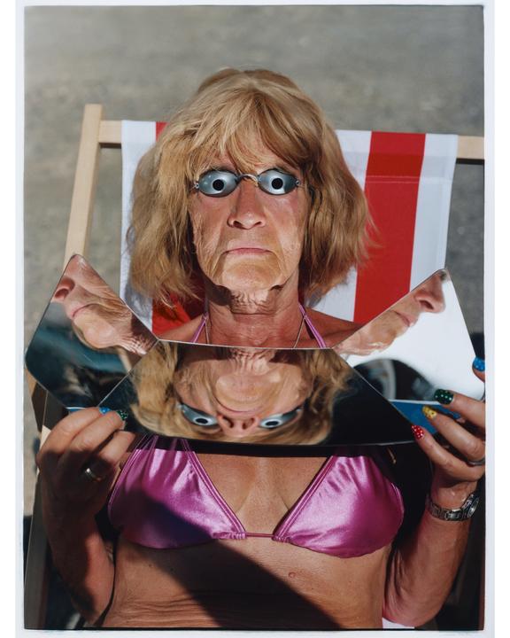 A woman wearing a pink bikini top and swimming goggles holds pieces of broken mirror reflecting her face.
