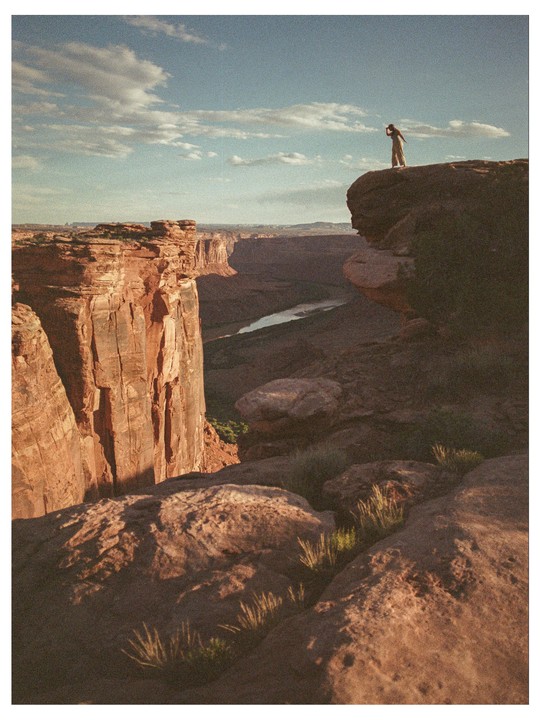 A woman stands on a cliff overlooking a canyon with a river winding through it under a partly cloudy sky.