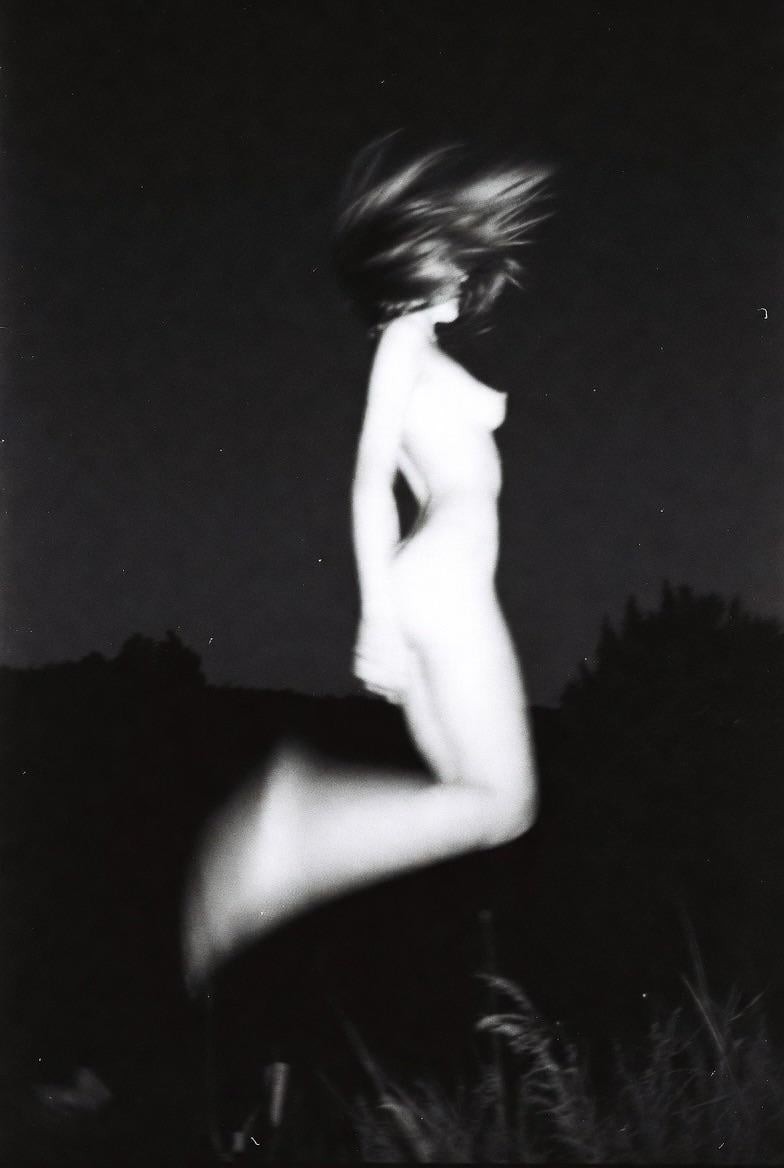 A blurry woman with long hair jumps in the dark with some foliage visible at the bottom of the frame.