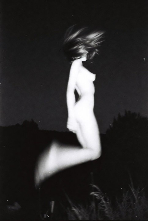 A blurry woman with long hair jumps in the dark with some foliage visible at the bottom of the frame.