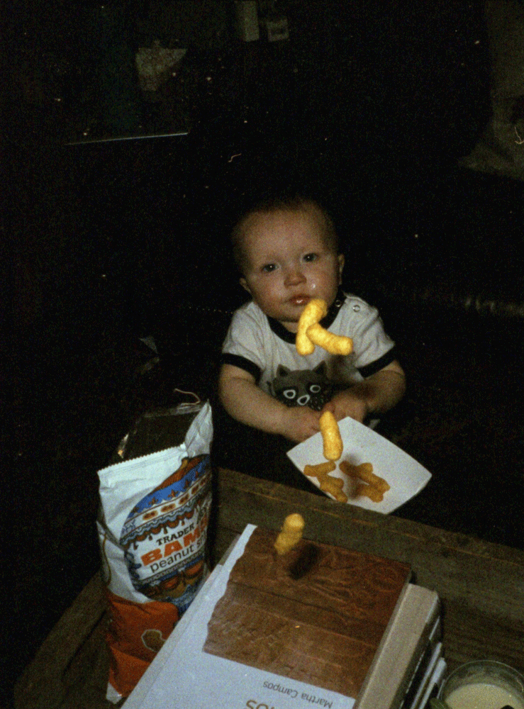 A child sits on the floor next to a bag of peanut puffs and a plate of snacks, with some snacks floating in the air.