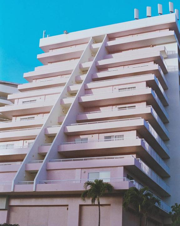 A pink building with balconies and a palm tree in the foreground stands against a bright blue sky.