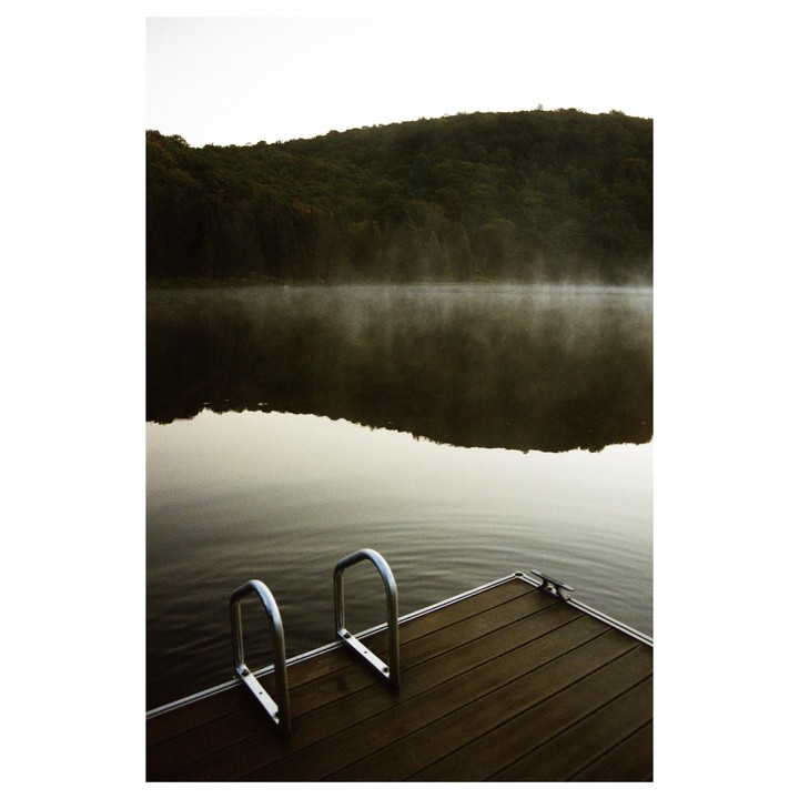 A wooden dock with metal handles extends into a misty lake, reflecting a tree-covered hill under a pale sky.