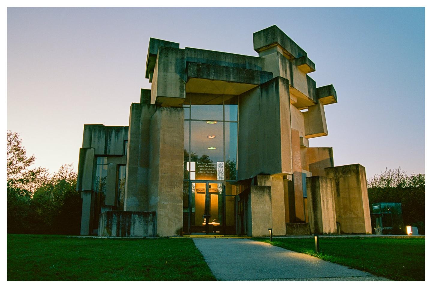 A brutalist church with geometric concrete forms stands against a twilight sky, with illuminated pathways leading to its entrance.