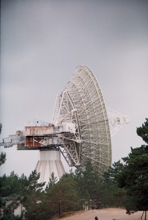 A large radio antenna stands above a forest and sandy ground under a grey sky.
