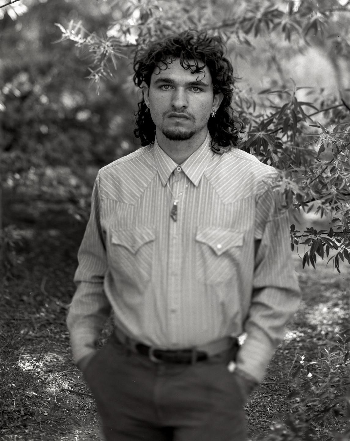 A man with curly hair and a beard stands outdoors wearing a striped shirt and a necklace.