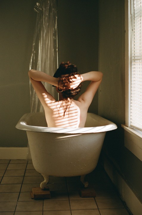 A woman sits in a bathtub with her back to the viewer, her hands behind her head, with shadows from a window blind cast across her back.