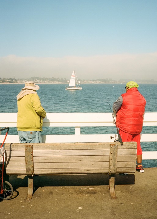 Two people stand on a pier, one fishing, while a sailboat passes on the hazy water in the background.