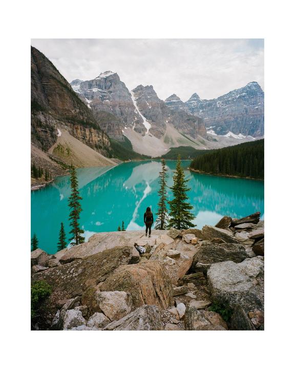 A woman stands on a rocky outcrop overlooking a turquoise lake surrounded by mountains and pine trees.
