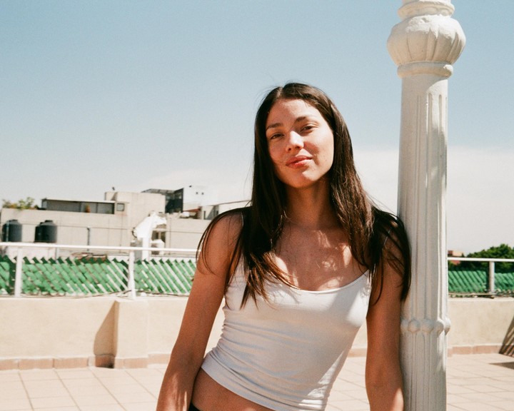 A woman with long hair leans against a white pillar on a rooftop.