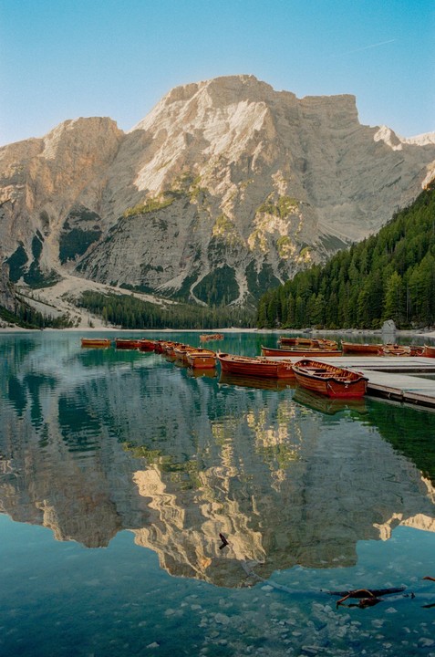 Wooden boats are moored to a dock on a clear lake with a large mountain and forest in the background.