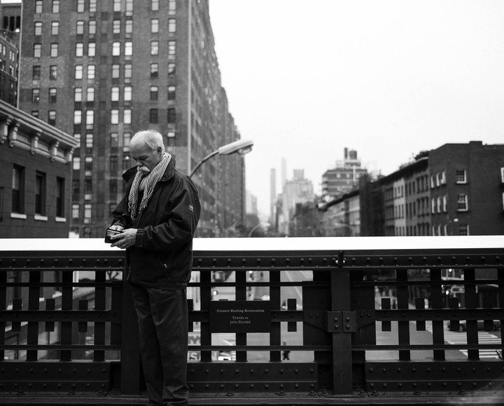 A man with a mustache and scarf stands on a bridge looking down at a small device in his hands, with city buildings in the background.