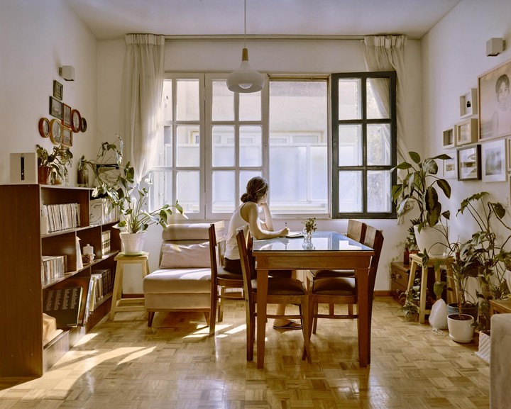 A woman sits at a dining table writing in a notebook, with plants and bookshelves surrounding her in a sunlit room.