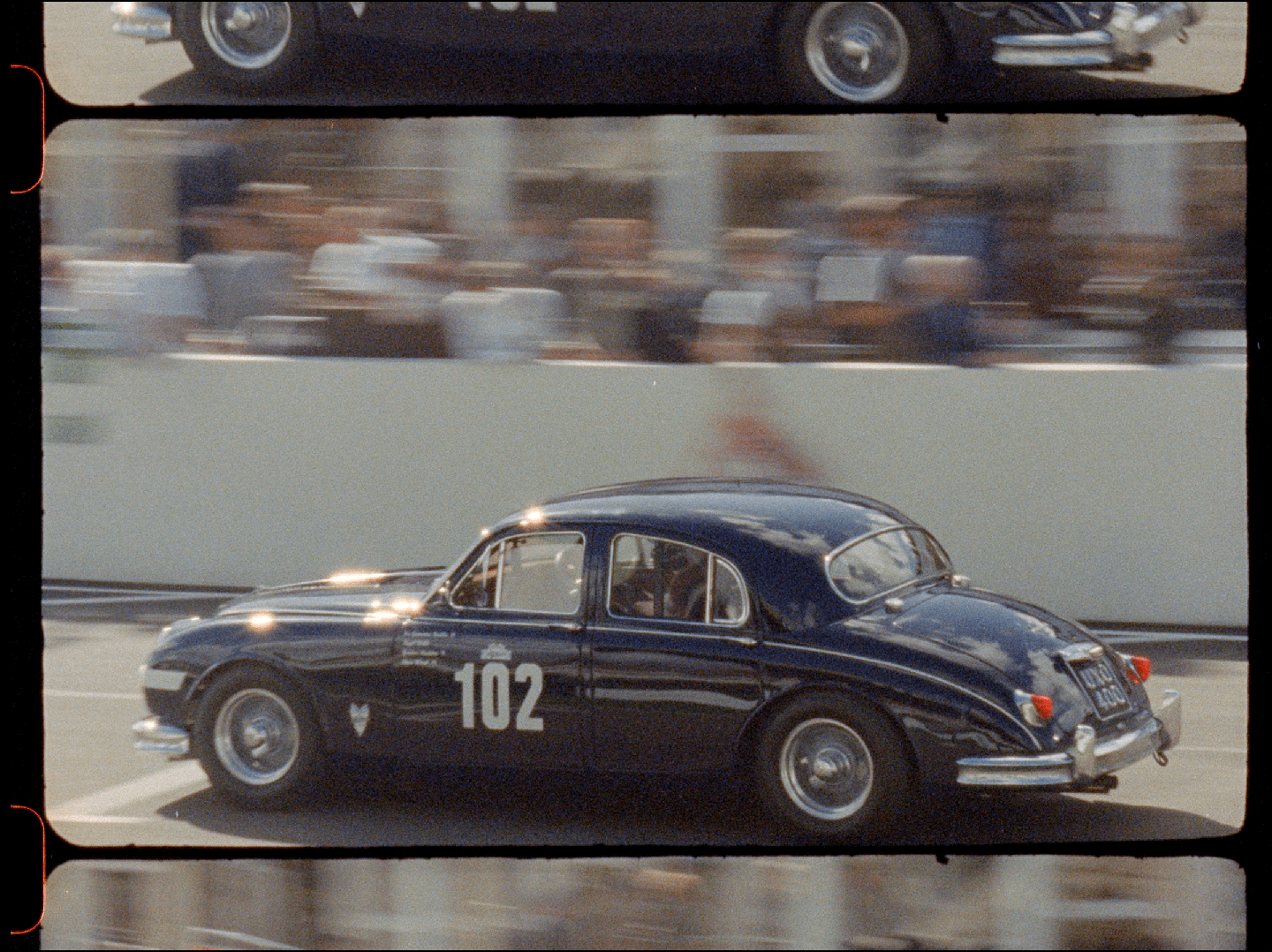 A dark blue vintage car with the number 102 on its side races past a blurred crowd.